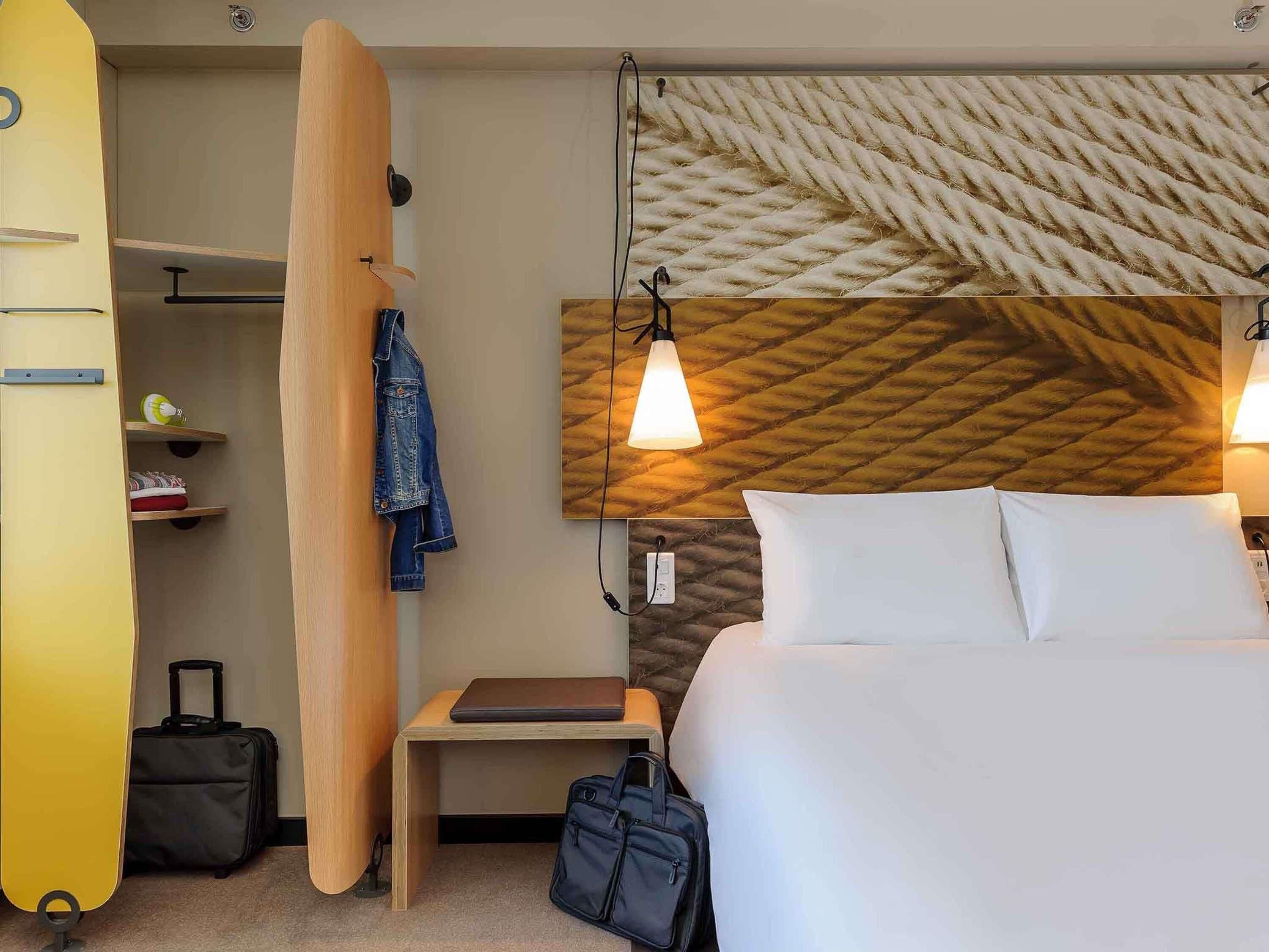 A hotel room with a bed, a desk, a surfboard, a suitcase, and a jacket hanging on the wall.