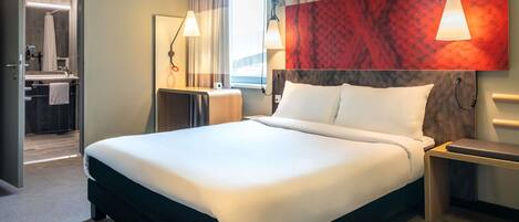 Quarto de hotel com uma cama grande, uma escrivaninha com um laptop, uma mesa de cabeceira, uma janela com cortinas, um banheiro com chuveiro e pia, e uma cabeceira com um desenho estampado em vermelho e cinza.