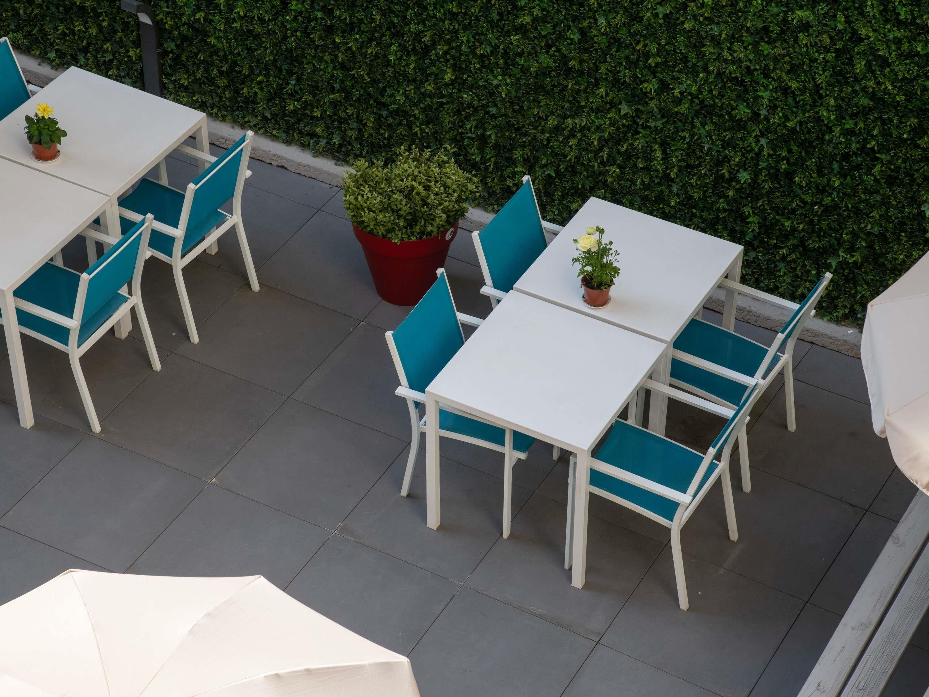 Un espace extérieur aménagé en terrasse, avec des tables blanches et des chaises bleues, entouré d’une haie verdoyante.
