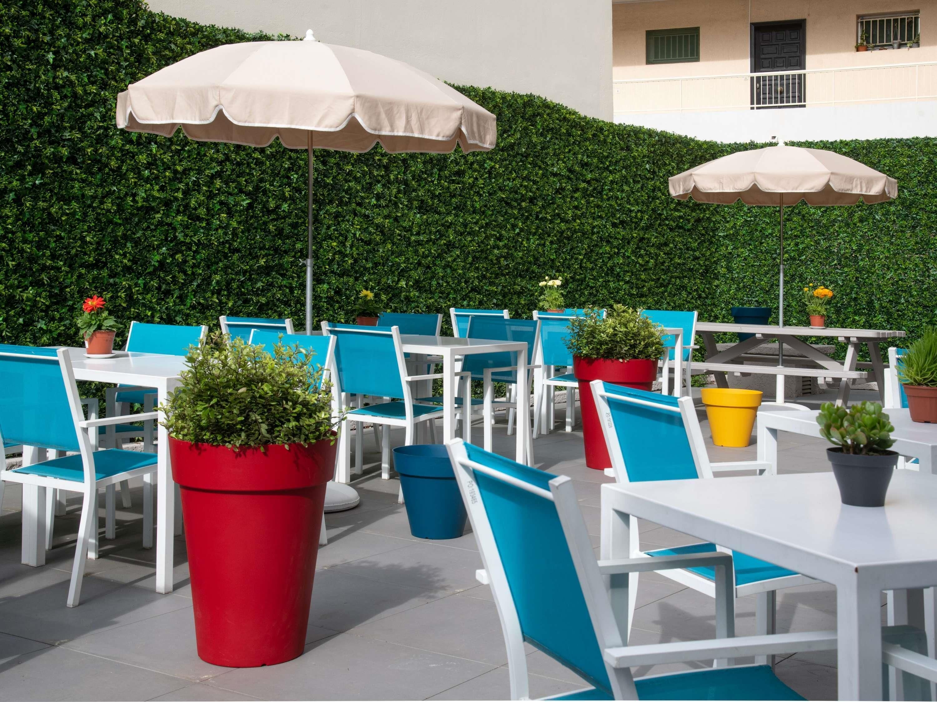 Een buitenzitplaats met witte tafels en blauwe stoelen, omgeven door groene hagen.