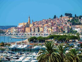 Une ville côtière aux bâtiments aux couleurs vives, un port de plaisance rempli de bateaux et un clocher d’église imposant.