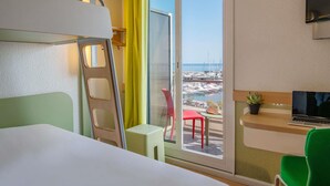 A hotel room with a bed, a desk with a laptop, a chair, a small table, and a view of a marina through a sliding glass door. - ibis budget Menton (Menton)