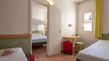 Une chambre d’hôtel avec un lit superposé, un bureau avec une chaise, un petit tabouret, une valise rouge et une fenêtre avec des rideaux jaunes.