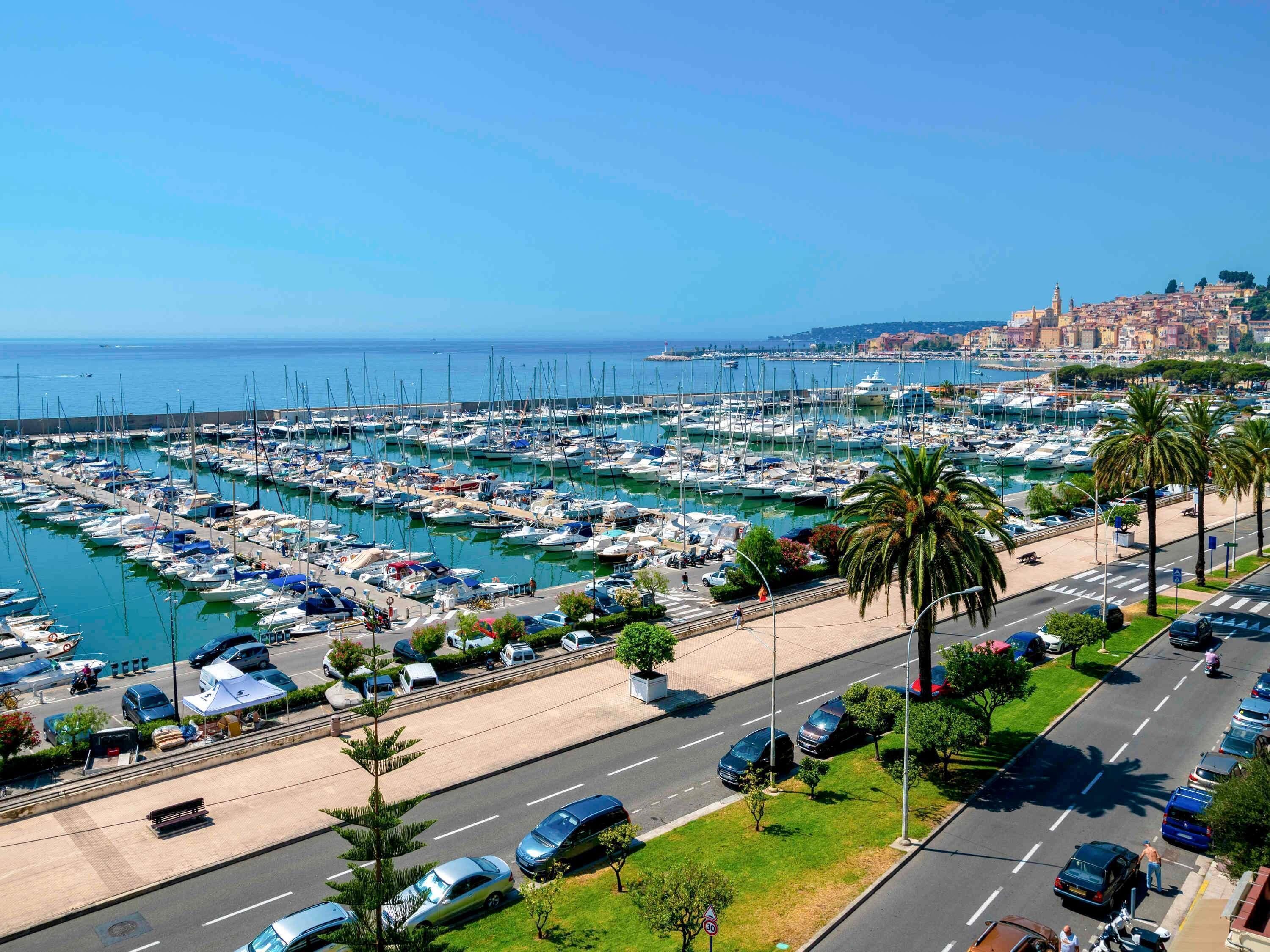 Un port de plaisance avec des bateaux amarrés, une route avec des voitures et un paysage urbain en arrière-plan.