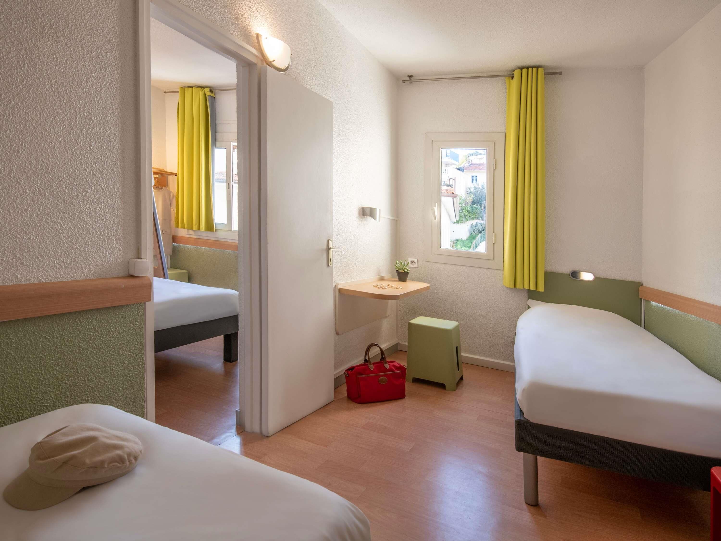 Ein Hotelzimmer mit zwei Betten, einem kleinen Tisch mit einer Pflanze, einer roten Tasche und einem Fenster mit gelben Vorhängen.
