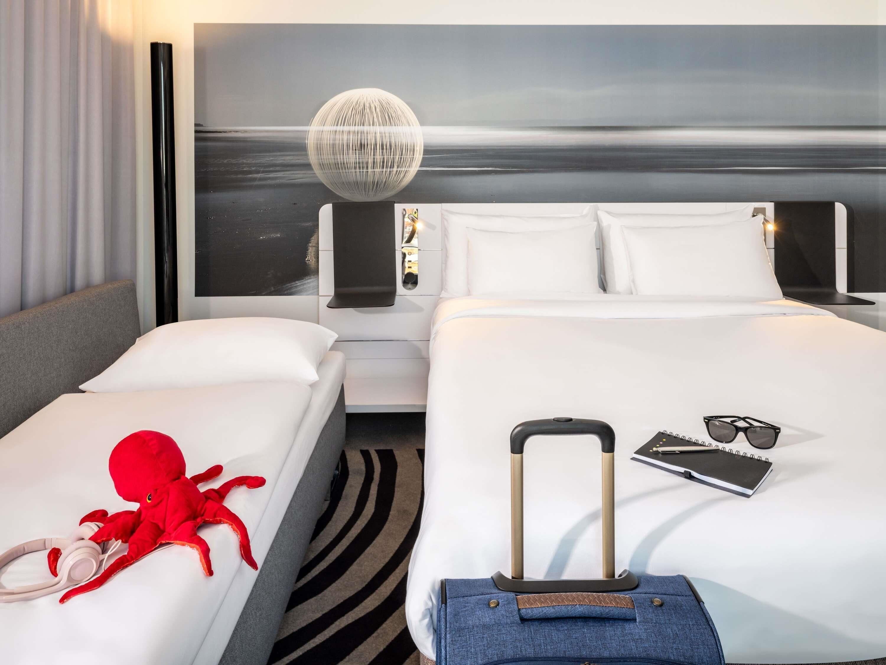 A hotel room with two beds, a large wall-mounted spherical light fixture, a black and white striped rug, and a suitcase on one bed.
