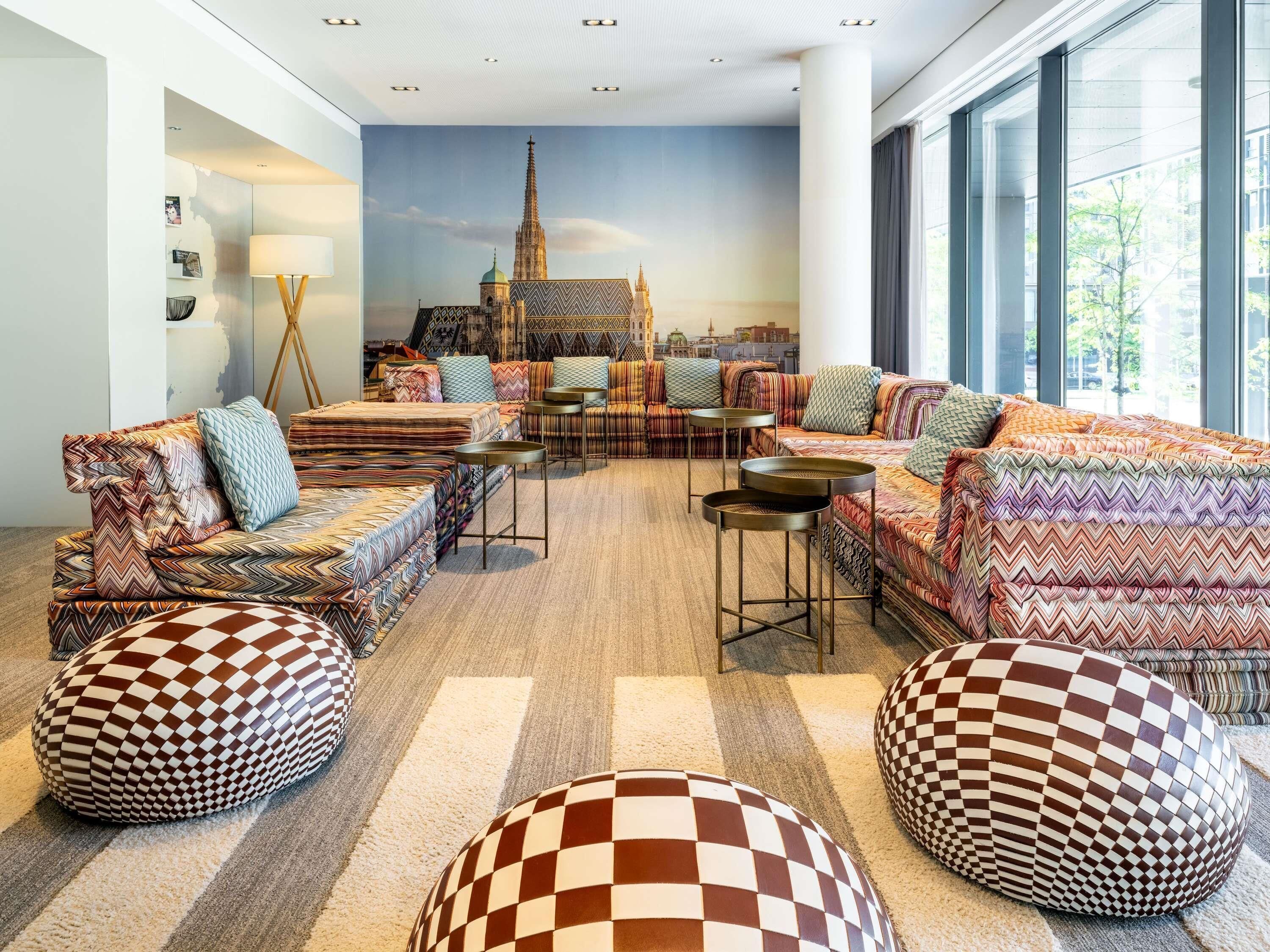 Ein modernes Wohnzimmer mit Stadtansicht-Wandgemälde, gemusterten Sofas, Hocker und einem großen Fenster.