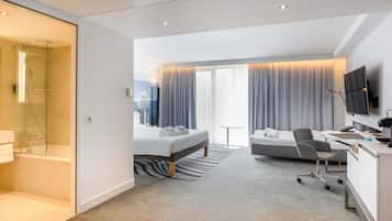 A modern hotel room with a glass-enclosed shower, a bed with a view, a desk with a chair, and a flat-screen TV.