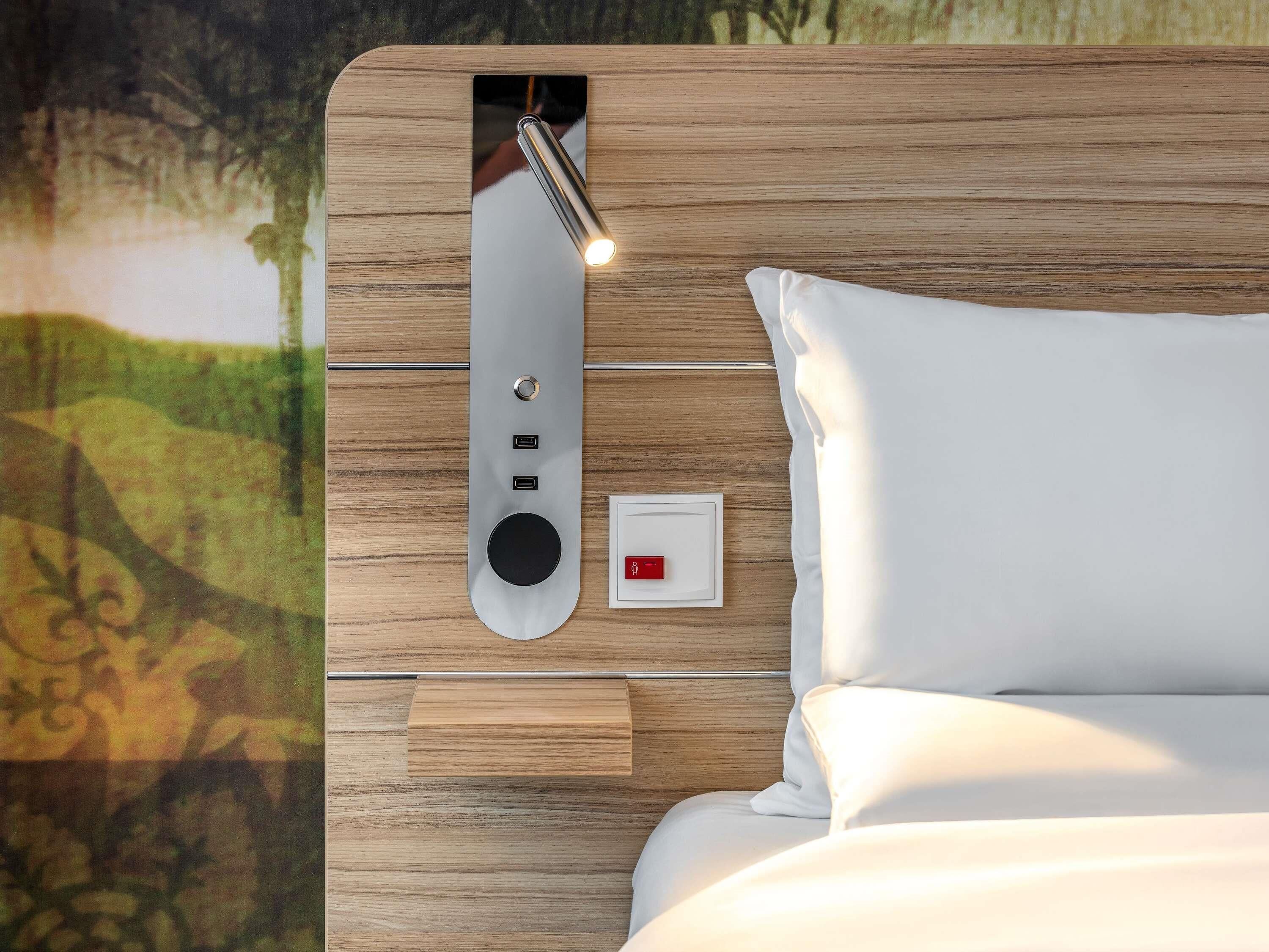 A modern hotel room with a wooden headboard, a bedside table, a nightstand, and a wall-mounted control panel with a red emergency light.