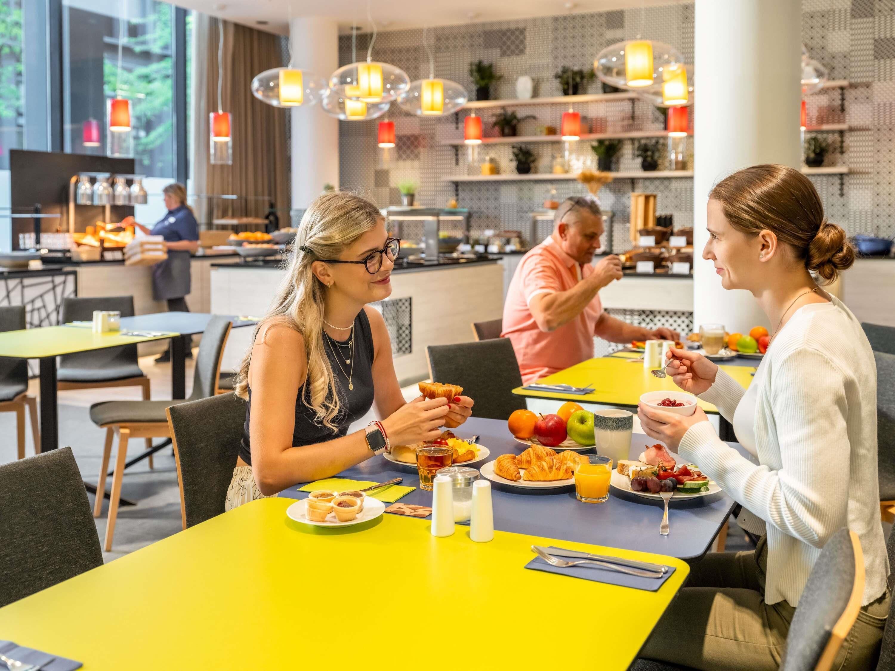Un comedor moderno con mesas amarillas, diversas opciones de desayuno y un barista preparando bebidas.