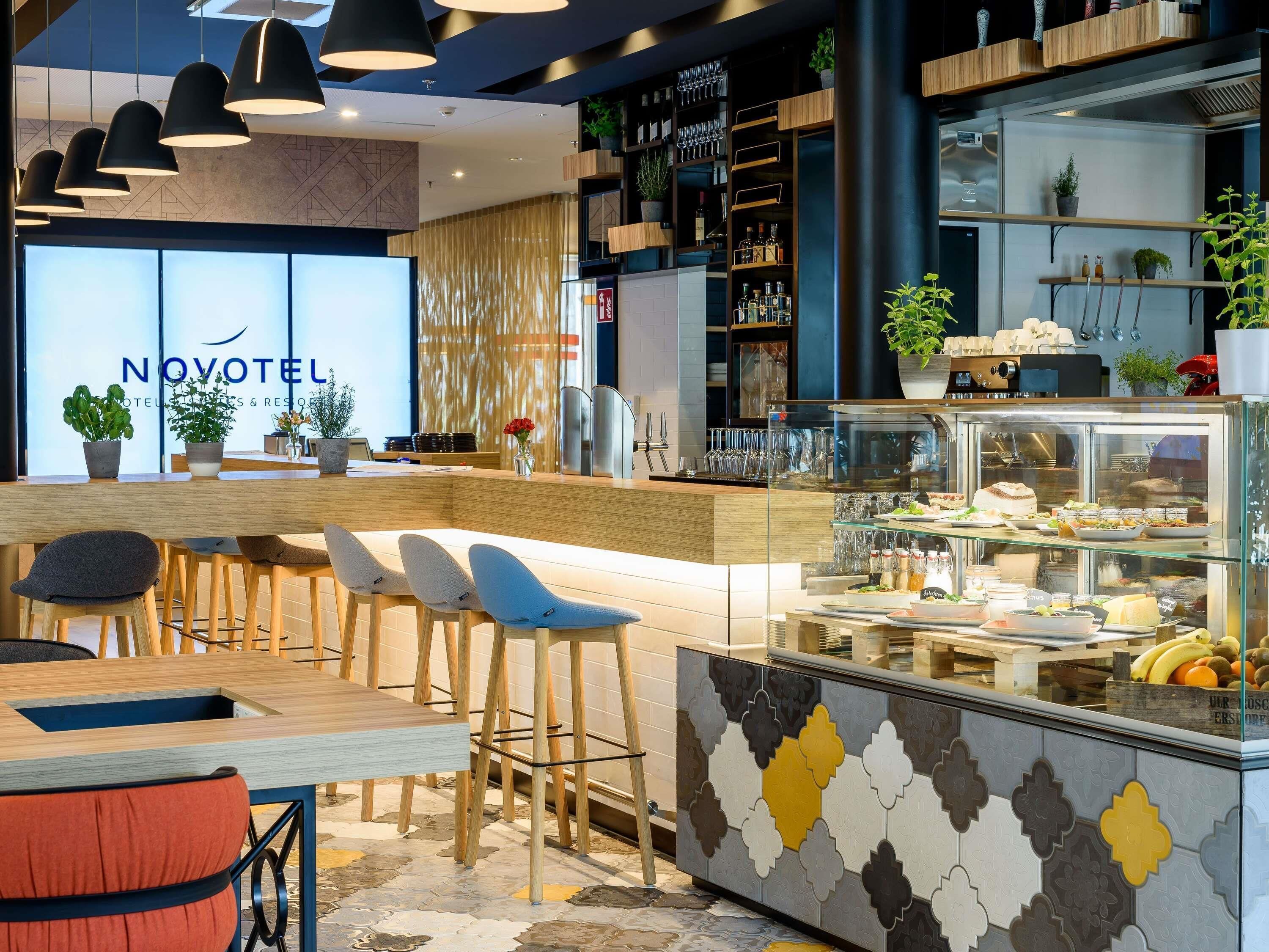 Bar moderno de hotel com balcão exibindo comida e bebidas, banquetas e área de recepção visível com a placa 'NOVOTEIL'.