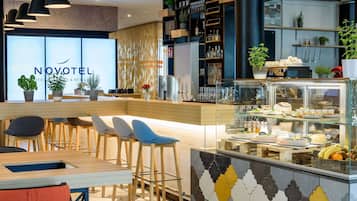 Bar moderno de hotel com balcão exibindo comida e bebidas, banquetas e área de recepção visível com a placa 'NOVOTEIL'.