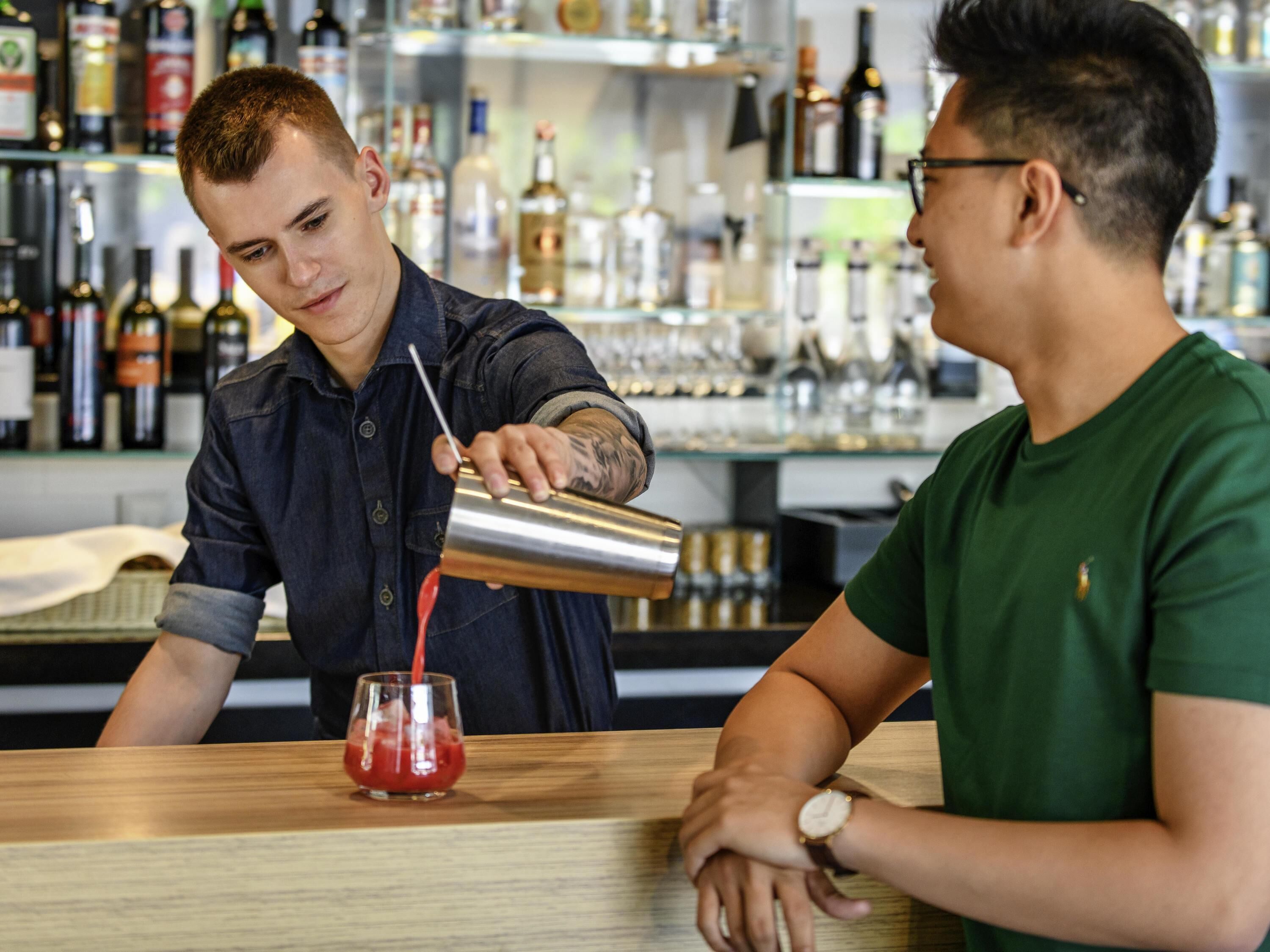 A bartender pouring a drink into a glass at a bar with shelves of bottles in the background.