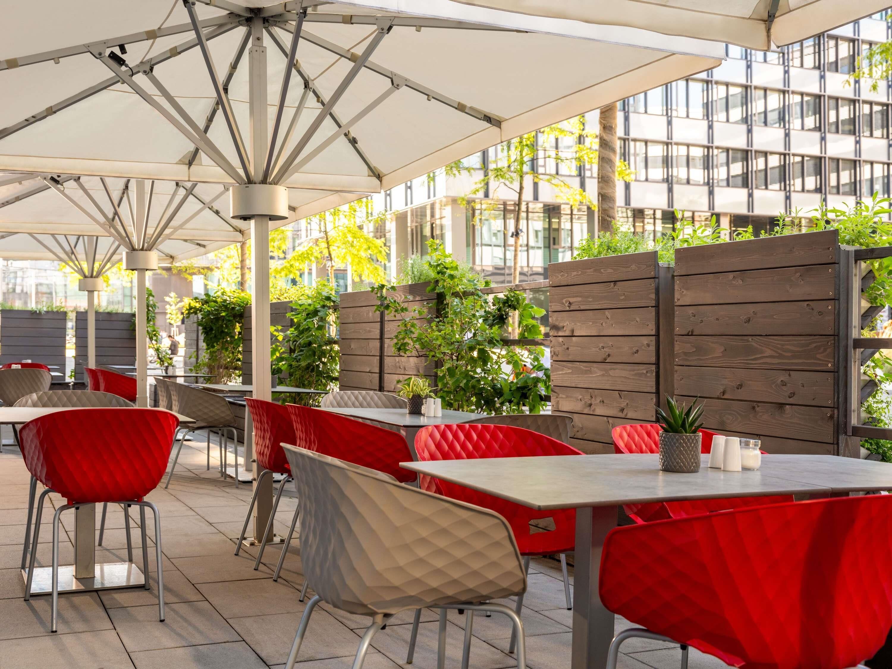 Un café al aire libre con sillas rojas y grises, mesas blancas y un separador de madera con plantas.