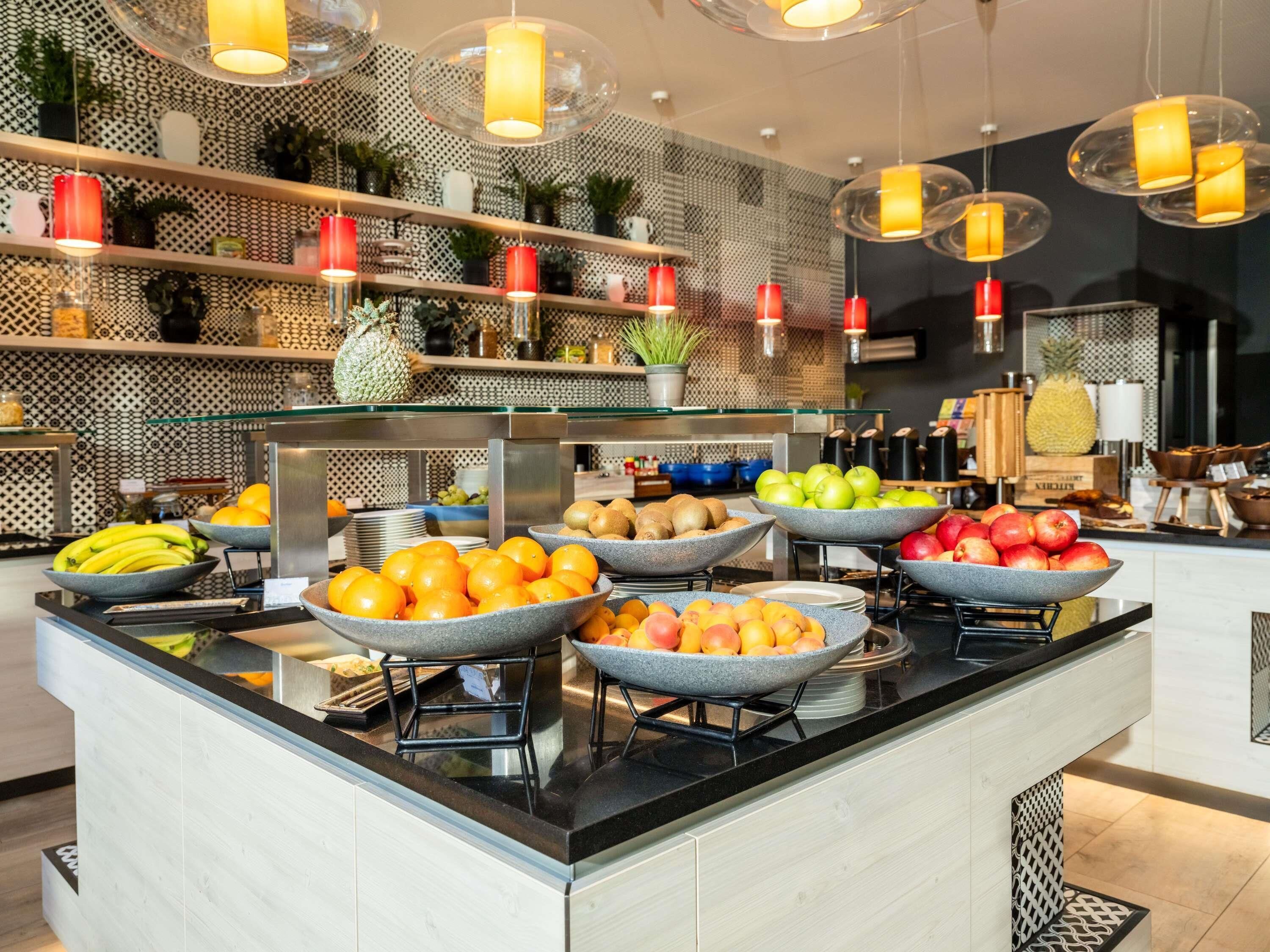 A buffet with various fruits and vegetables, a modern kitchen area, and hanging pendant lights.