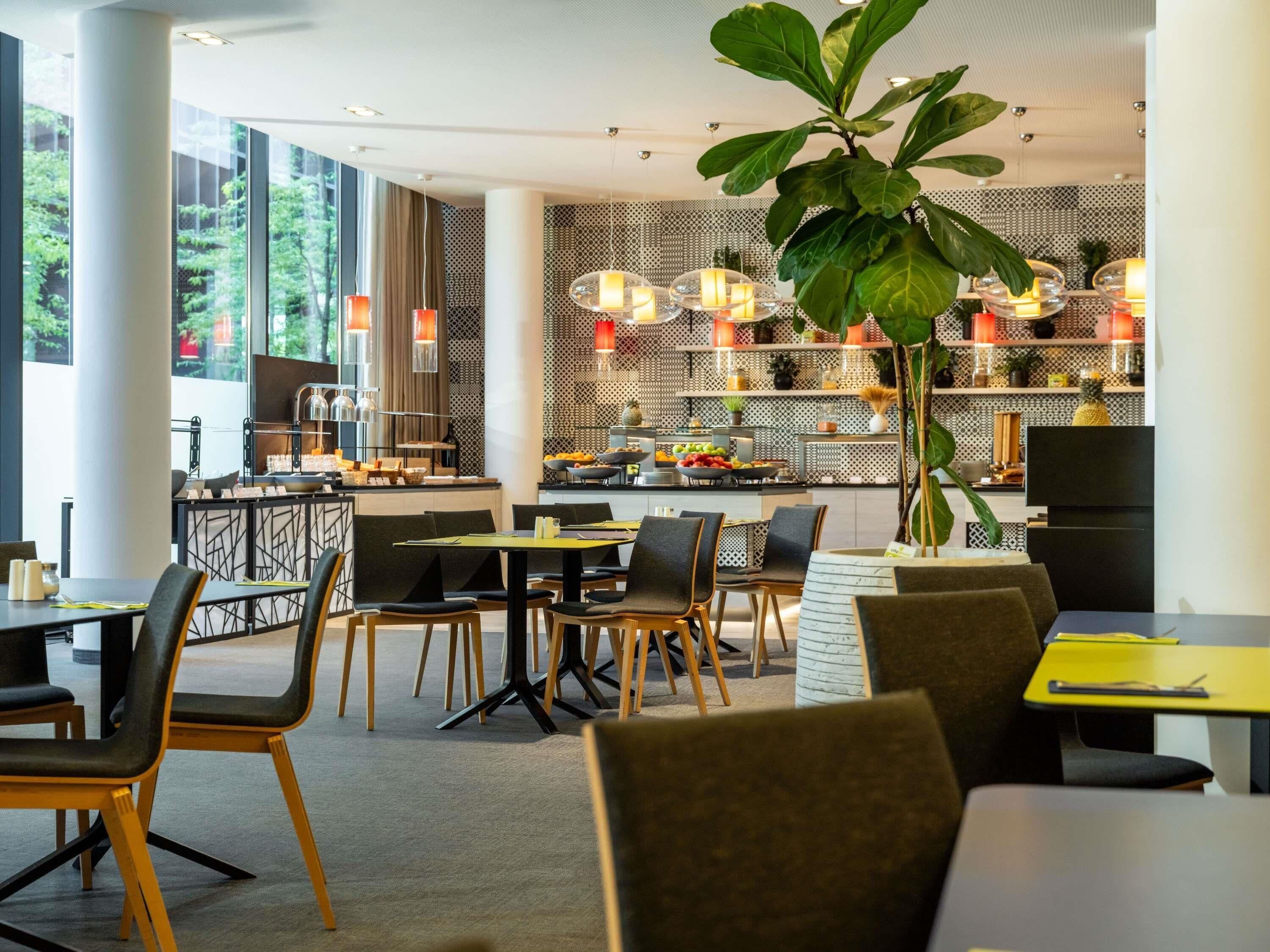 Un espace repas moderne avec des tables et des chaises, une grande plante et une cuisine en arrière-plan.