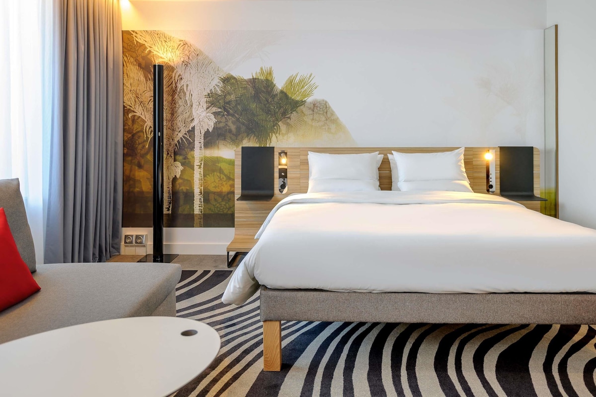 Una habitación de hotel con cama, mesita de noche, lámpara, silla y un mural en la pared.