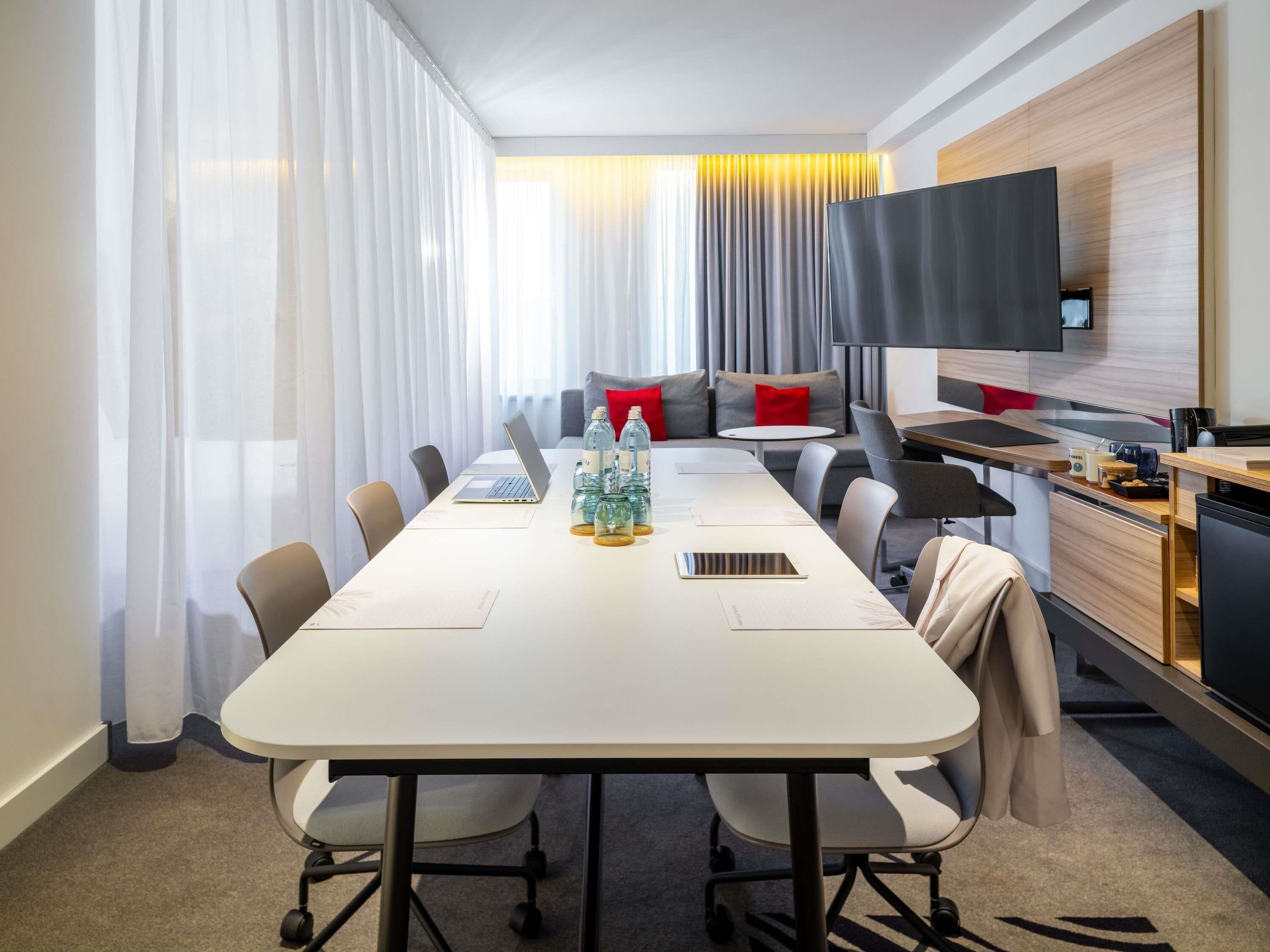 A modern meeting room with a long table, chairs, a sofa with cushions, a flat-screen TV, and a kitchenette area.