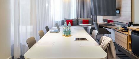 A modern meeting room with a long table, chairs, a sofa with cushions, a flat-screen TV, and a kitchenette area.
