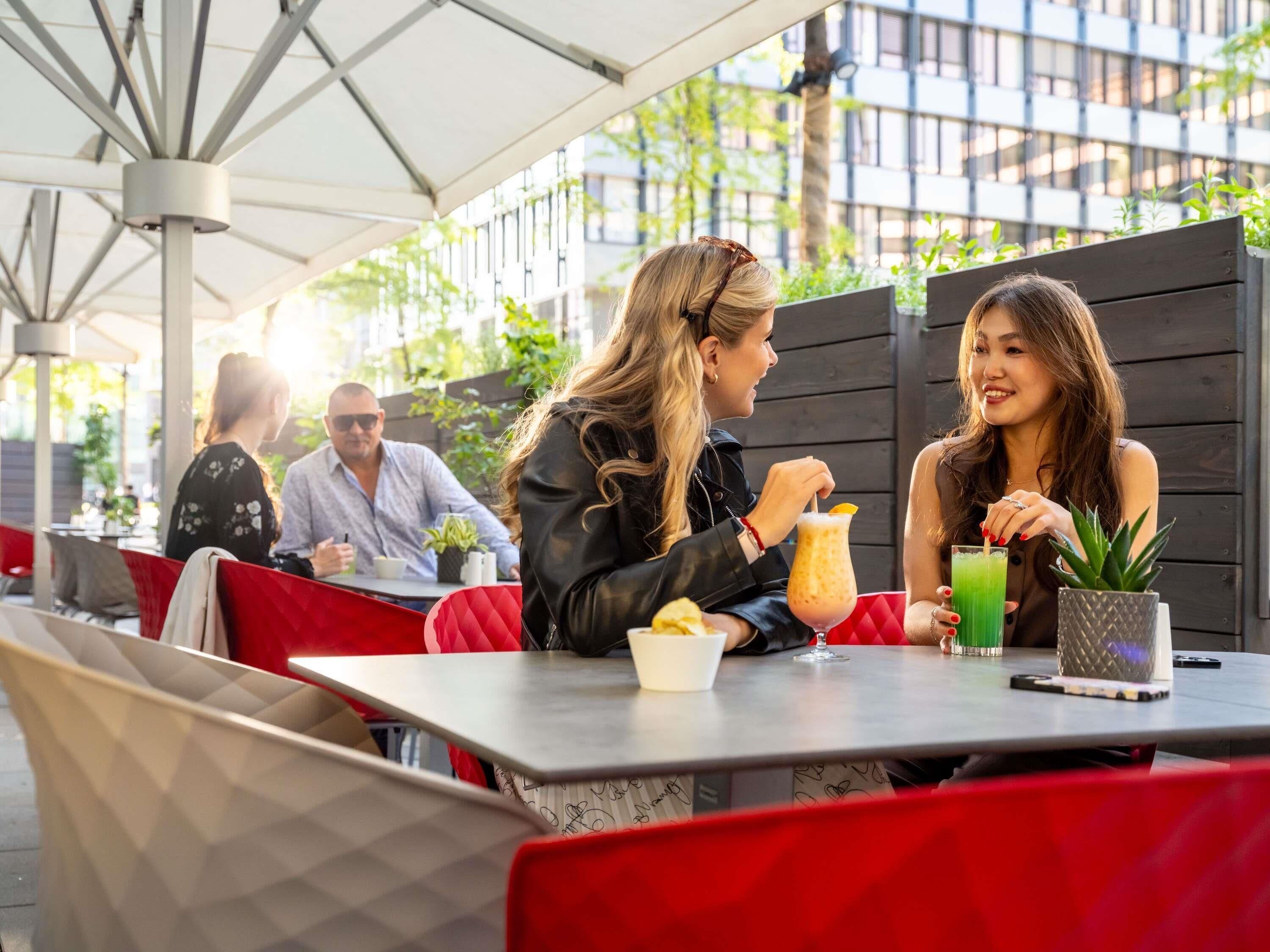 An outdoor café with tables and chairs, a person drinking a beverage, and another person eating.