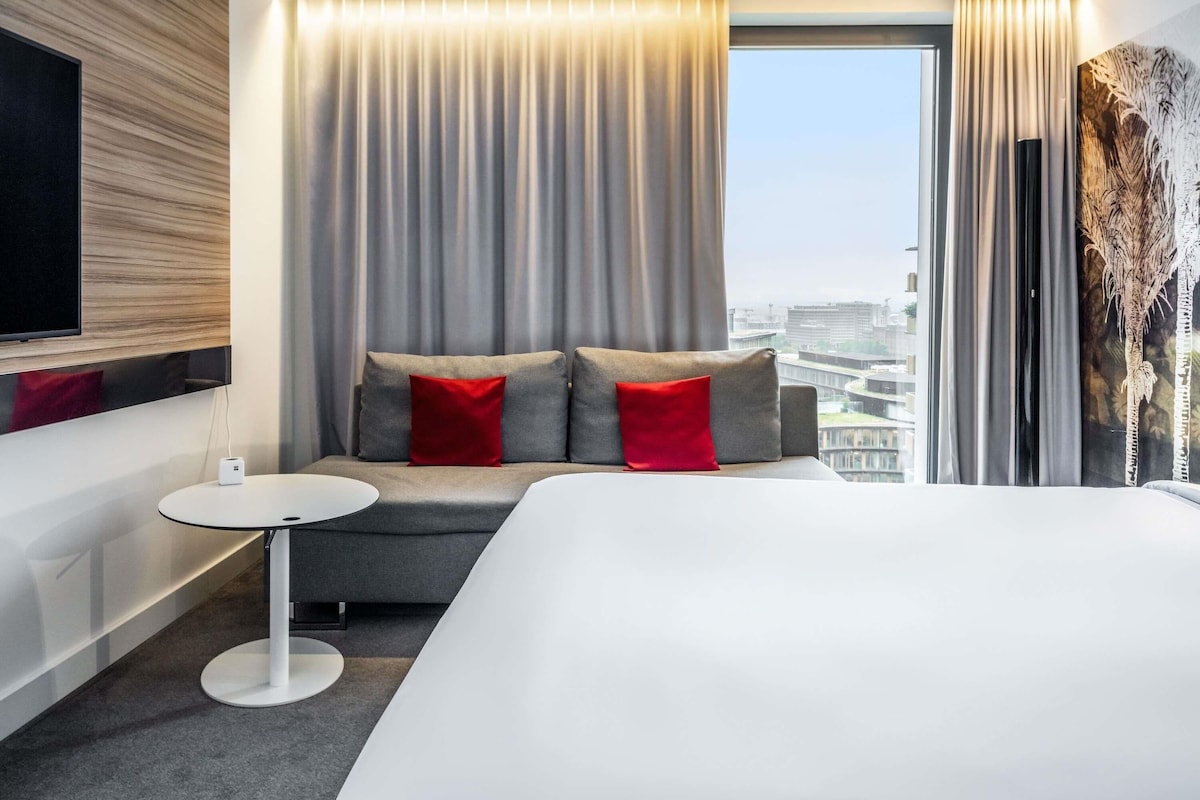 Una habitación de hotel moderna con una cama grande, un sofá con cojines rojos, una mesita redonda y un televisor montado en la pared.