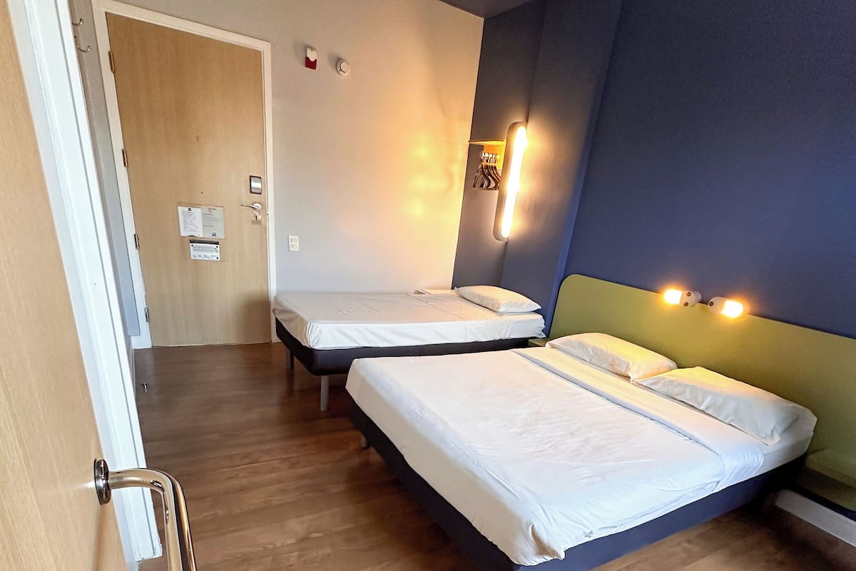 Quarto de hotel com duas camas, piso de madeira, porta com corrimão e luminária fixada na parede.