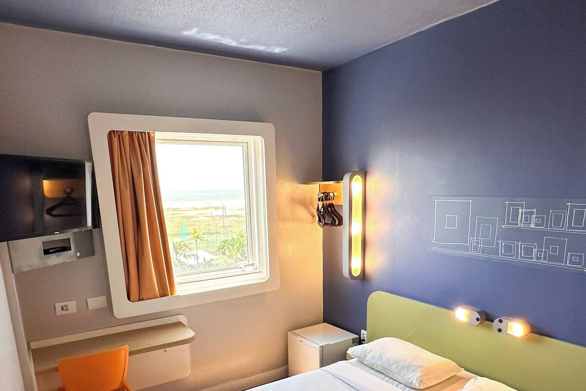 Quarto de hotel com cama, escrivaninha, cadeira, mesa de cabeceira, abajur e janela com cortinas.