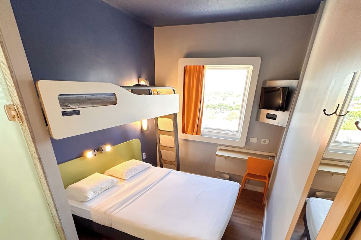 Quarto compacto com beliche, escrivaninha, cadeira e janela com cortinas.