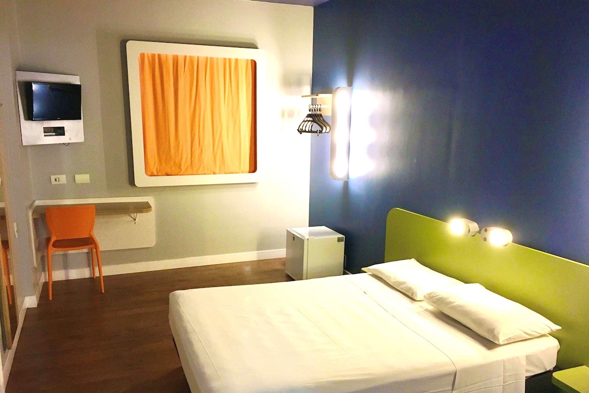 Habitación de hotel con cama, escritorio, silla, televisor y ventana con cortinas.