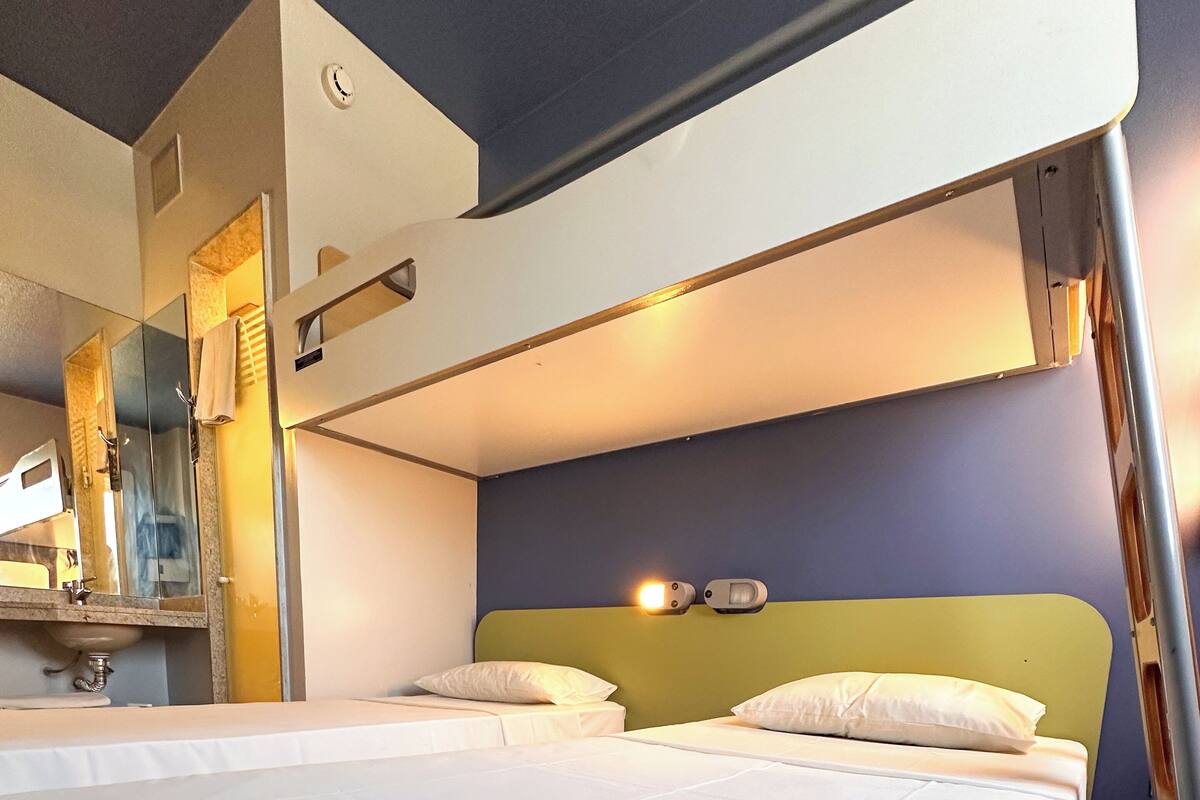 Quarto de hotel com duas camas, banheiro com espelho e pia.