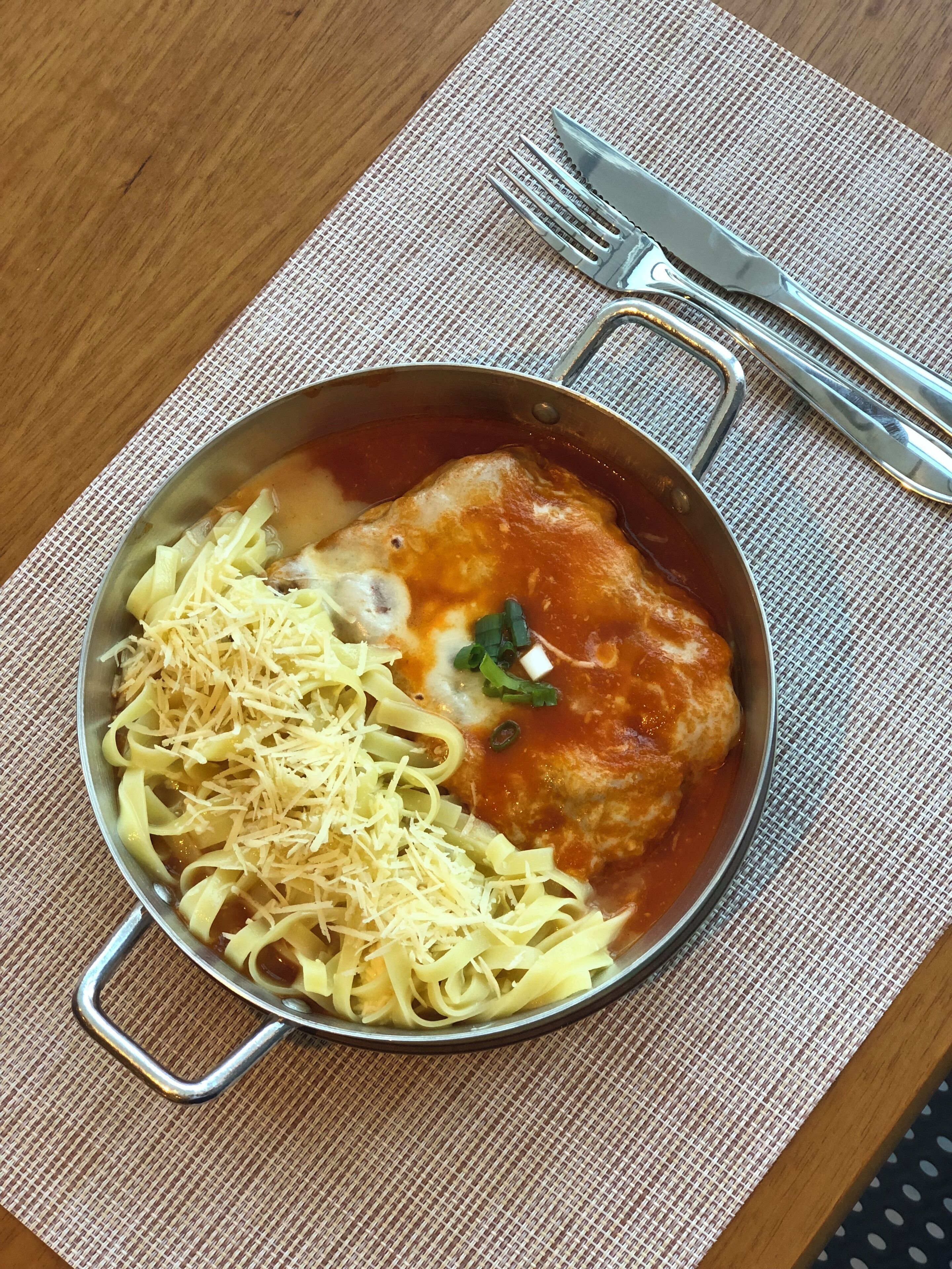 A bowl of food with noodles, shredded cheese, and a piece of meat in a tomato-based sauce, placed on a woven placemat.