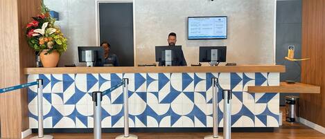Un mostrador de recepción con tres monitores de computadora, una pantalla digital que muestra información y una planta decorativa.
