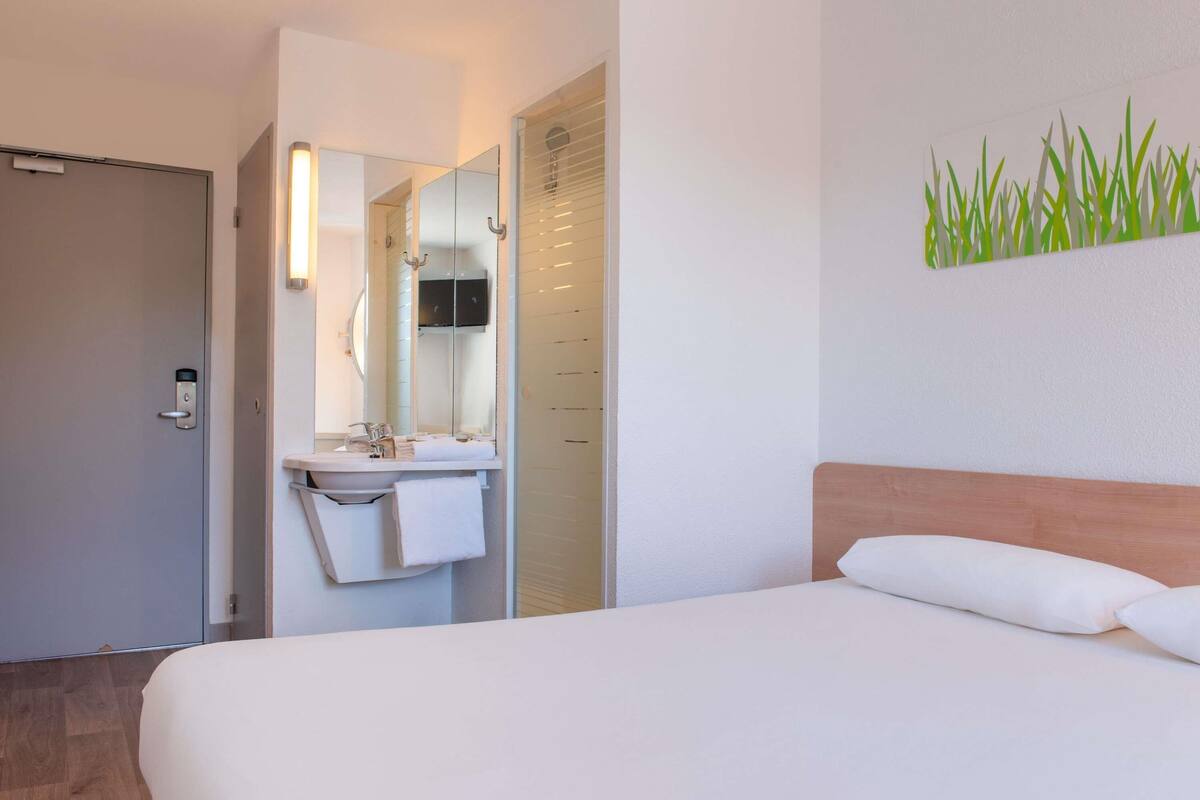 A hotel room with a bed, a bathroom with a sink and mirror, and a wall-mounted artwork.