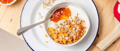 A bowl of cereal with milk, a spoon, and a small bowl of jam on a white plate with a blue rim.