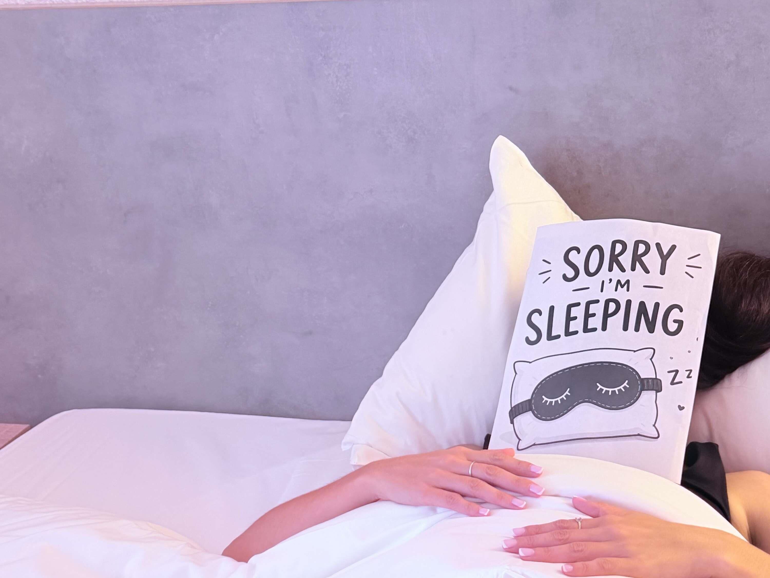 A person lying in bed with a pillow that has a drawing of a sleeping face and the text 'SORRY I'M SLEEPING'.