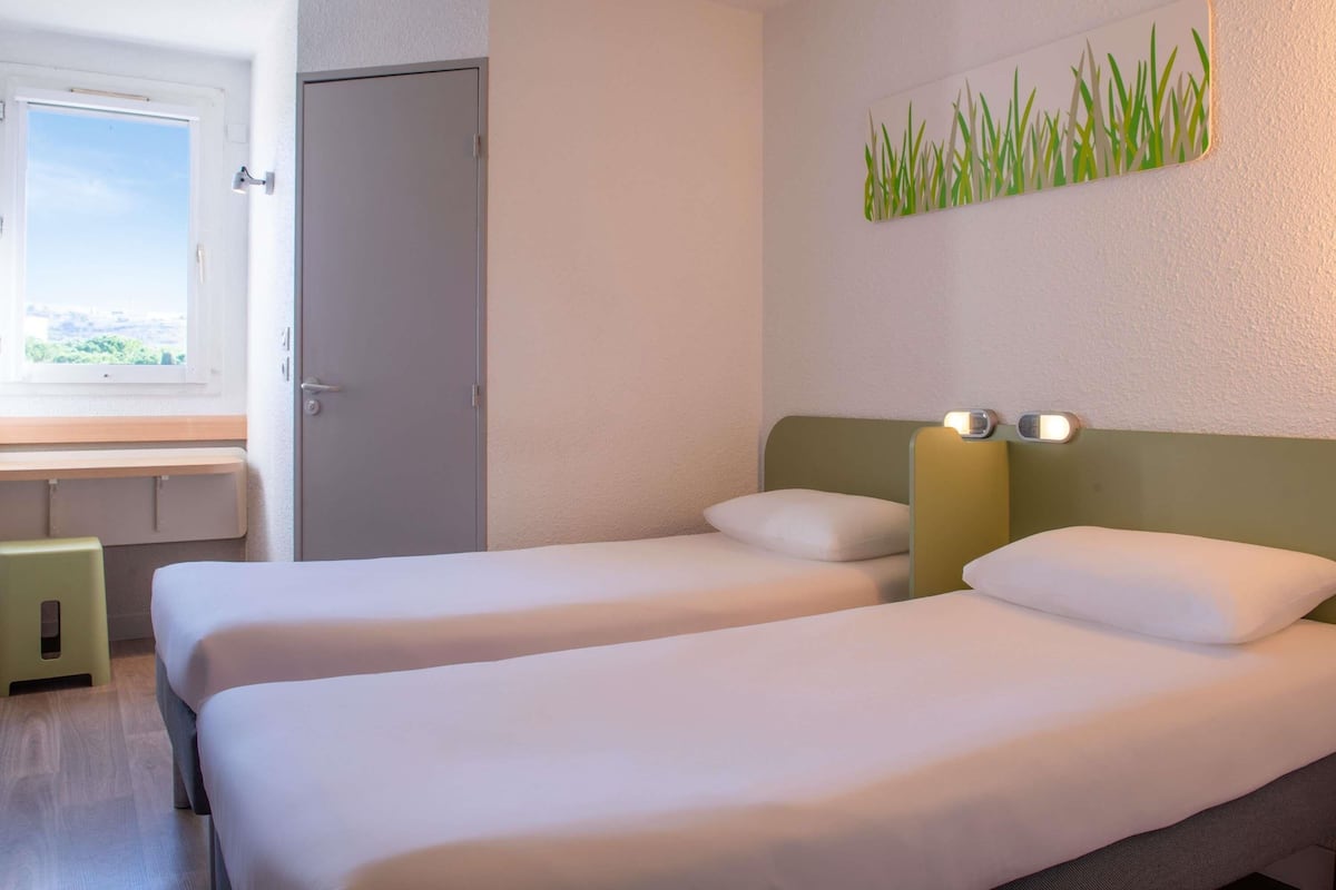 Habitación de hotel con dos camas individuales, una ventana con vistas, una puerta y una obra de arte colgada en la pared.