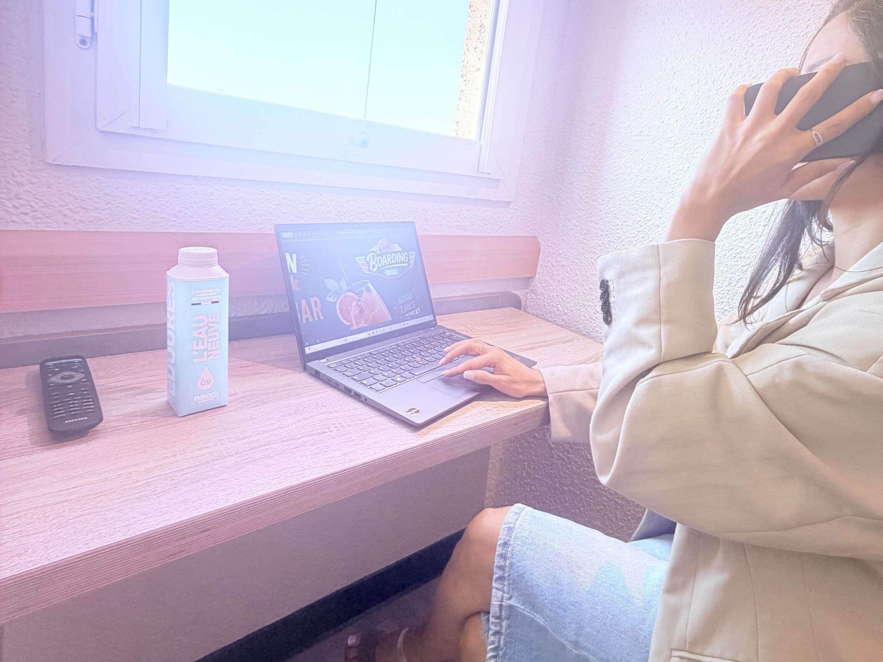 A person using a laptop on a wooden desk with a bottle of water and a remote control beside it.