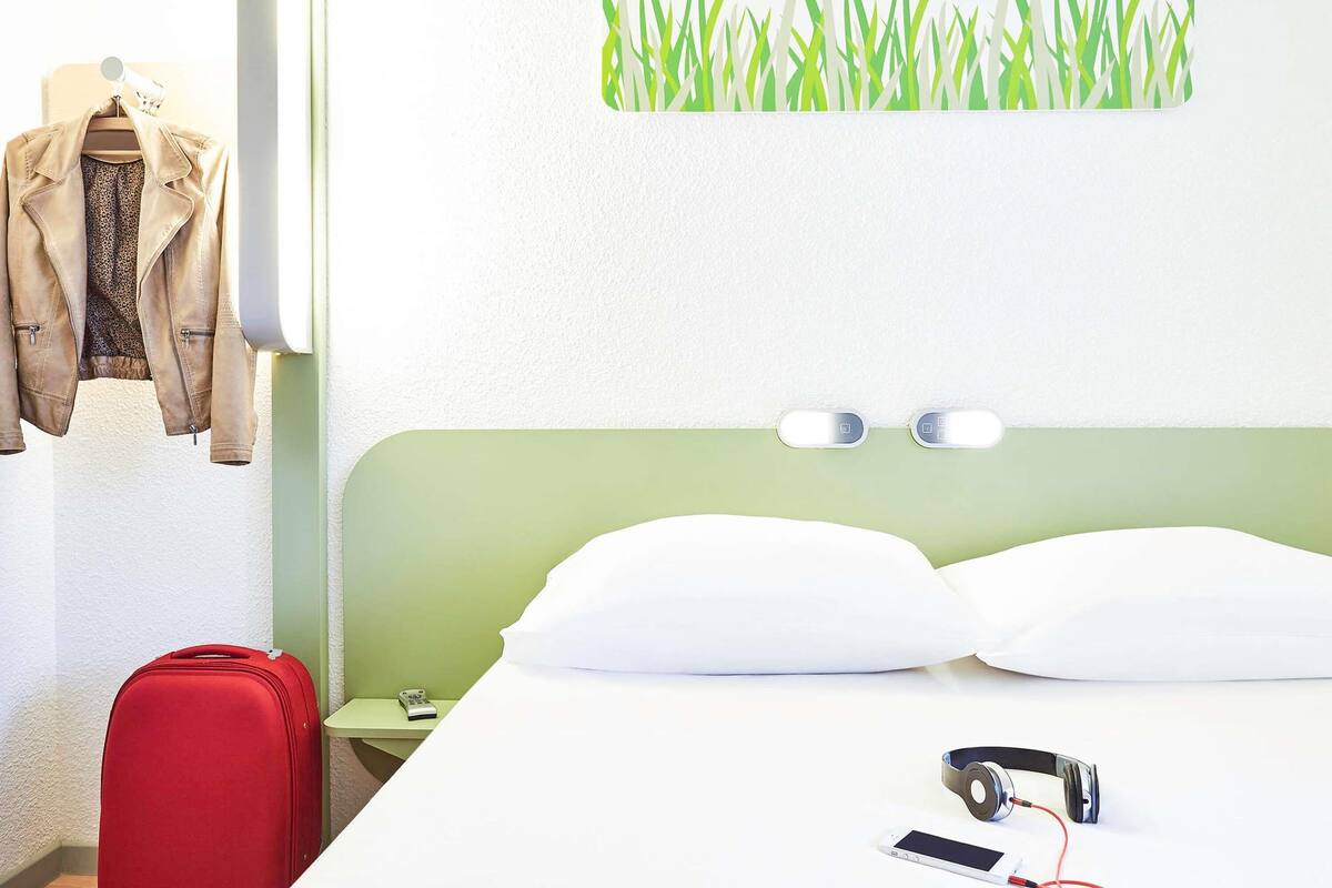 Habitación de hotel con cama, almohadas, una mesita de noche, una maleta roja, una chaqueta colgada de un perchero y una pared con diseño floral.