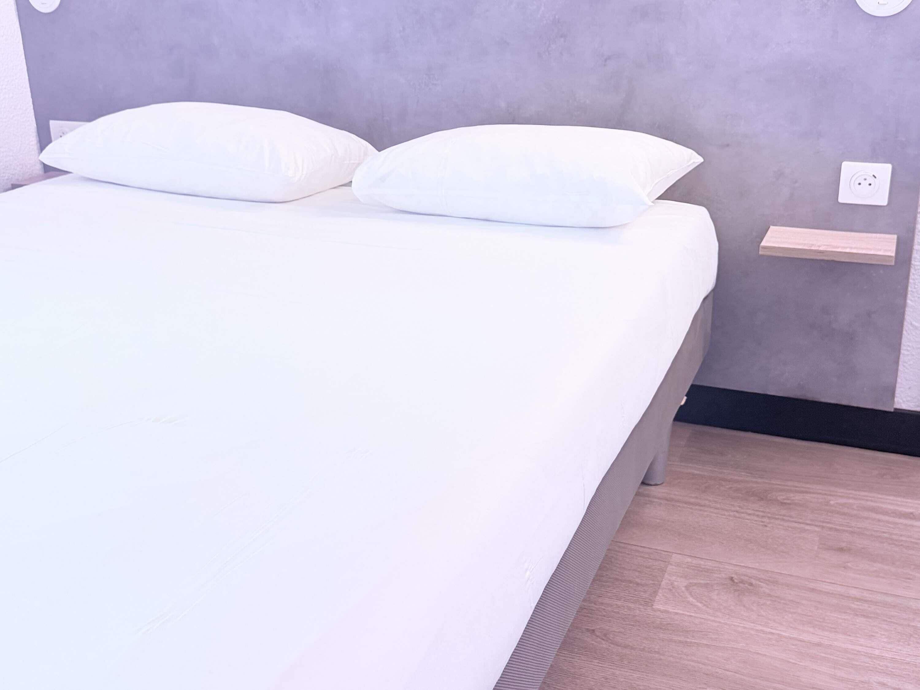 Un lit avec une literie et des oreillers blancs, un mur gris et un plancher en bois.