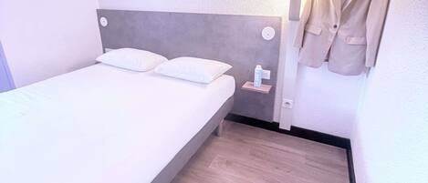 A single bed with white bedding in a room with a grey headboard. A small shelf with a bottle is mounted on the wall beside the bed.