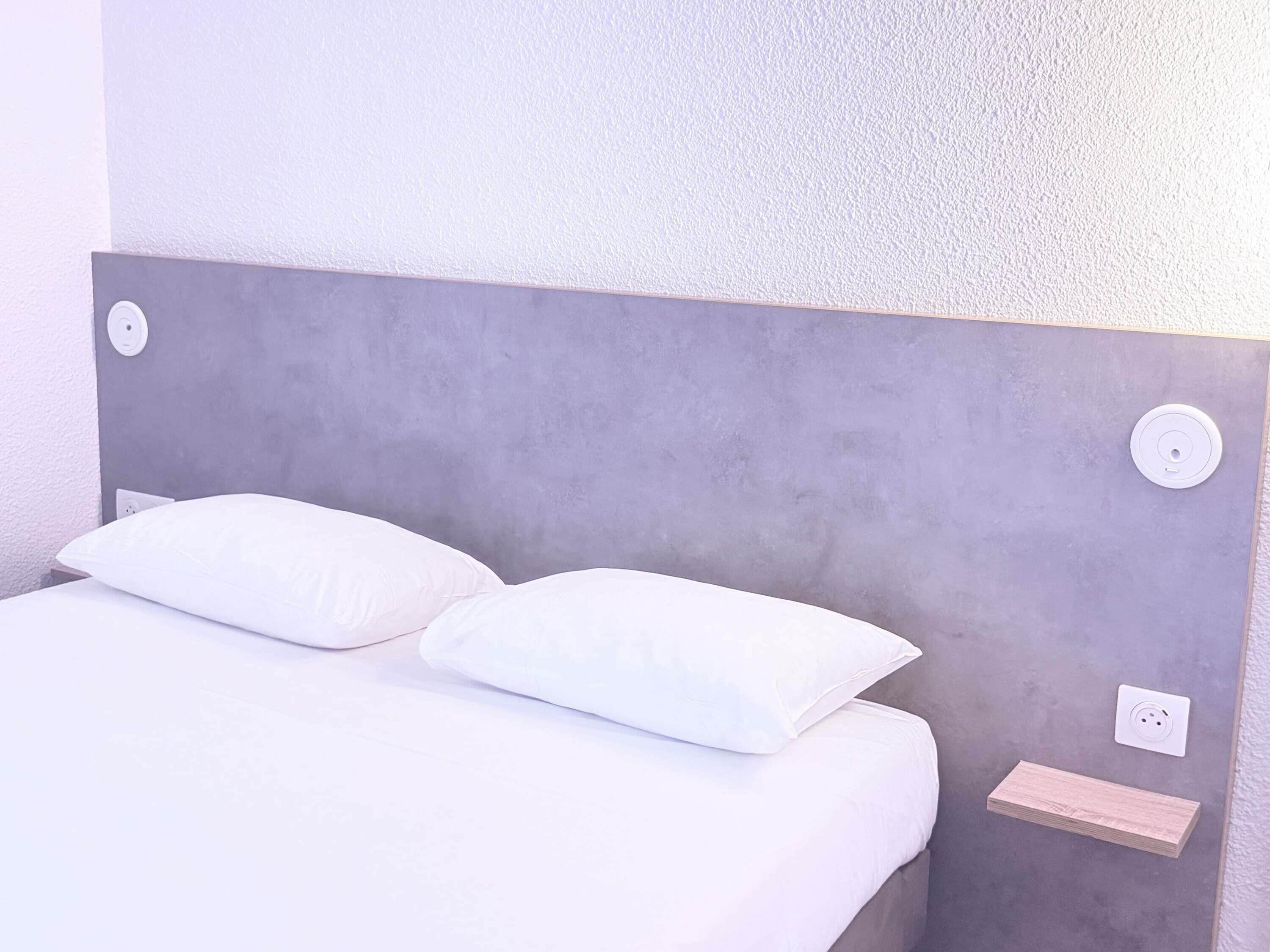 A bed with white bedding and pillows, a grey headboard, a bedside table with a light switch, and a power outlet.