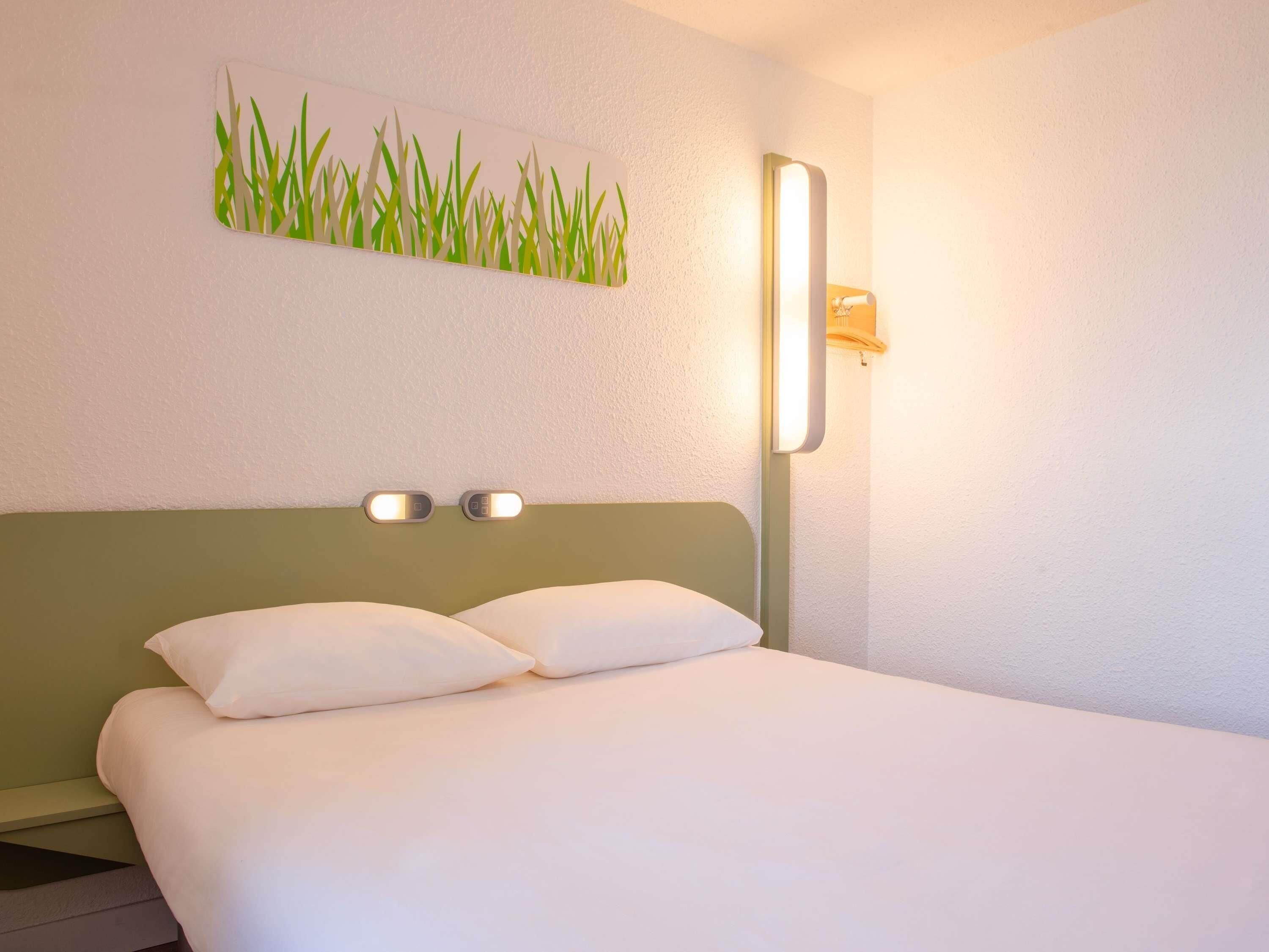 A hotel room with a bed, two bedside tables, a wall-mounted light, and a wall art piece.