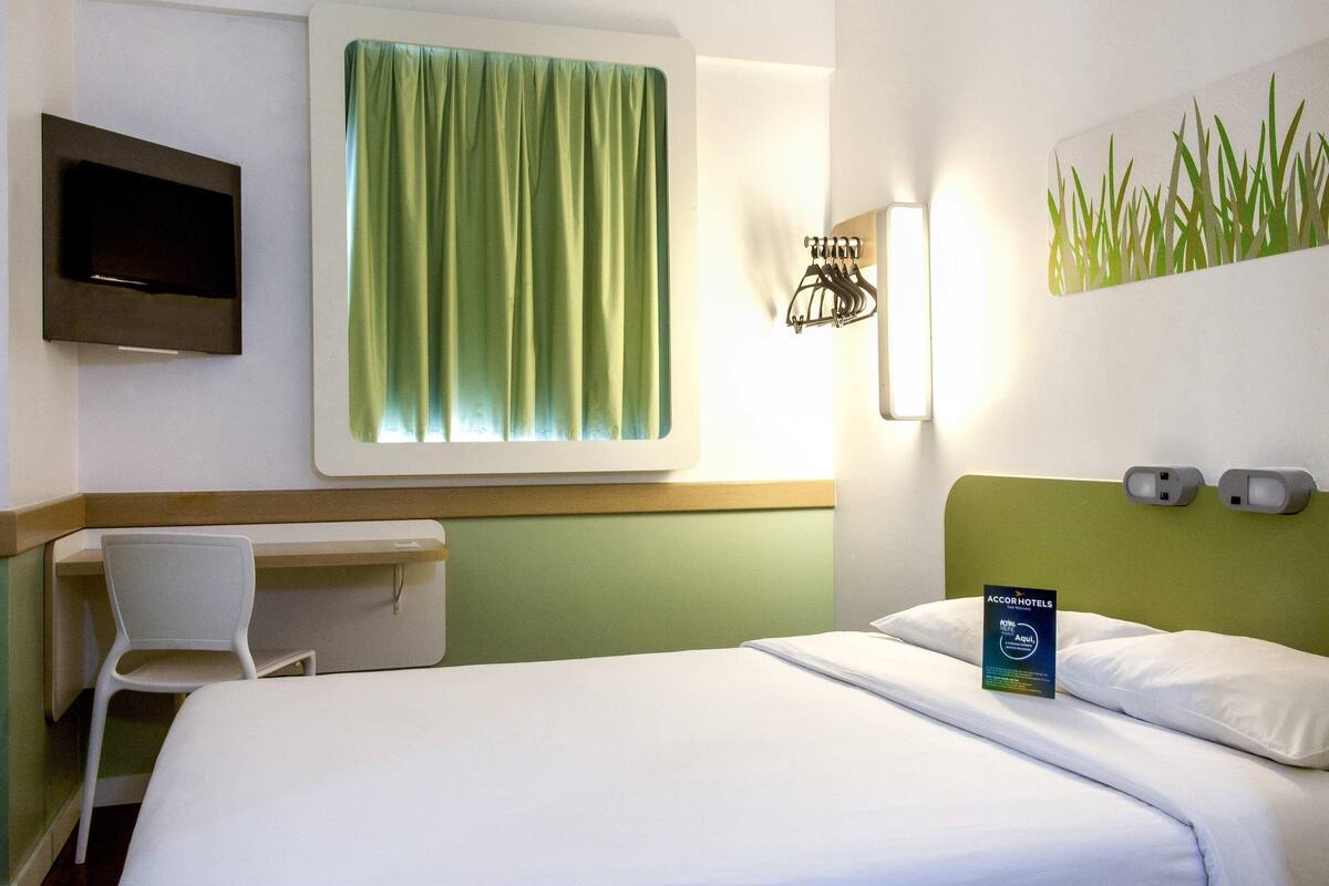 A hotel room with a bed, a desk, a chair, a television, and a wall-mounted light fixture.
