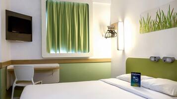 Quarto de hotel com cama, escrivaninha, cadeira, televisão e luminária fixada na parede.