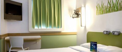 A hotel room with a bed, a desk, a chair, a television, and a wall-mounted light fixture.