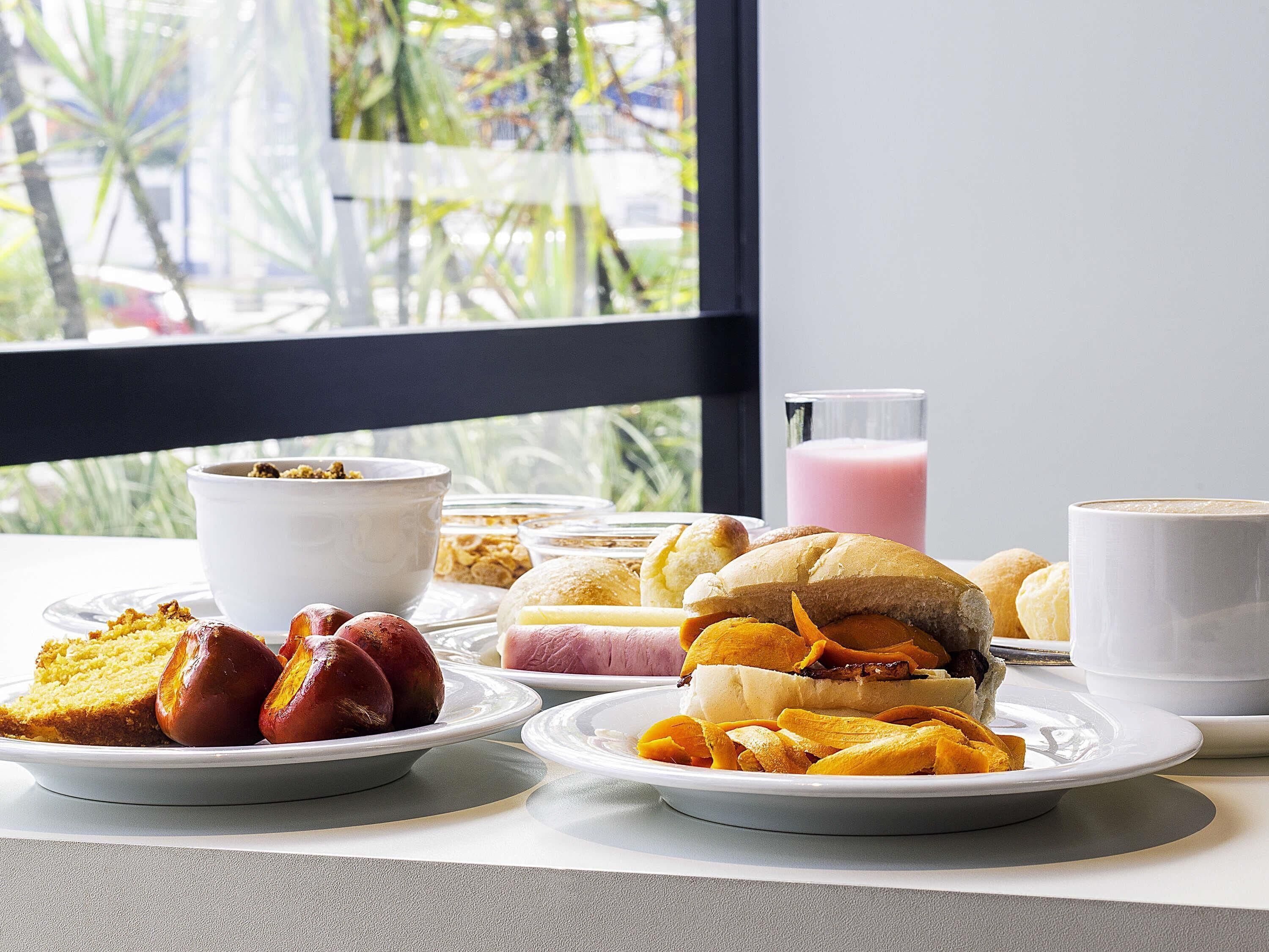 Disposição de café da manhã com diversos alimentos e uma bebida sobre uma mesa perto de uma janela.