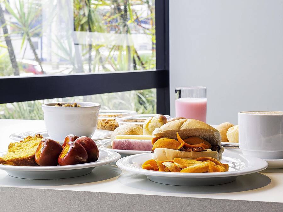 Disposição de café da manhã com diversos alimentos e uma bebida sobre uma mesa perto de uma janela.