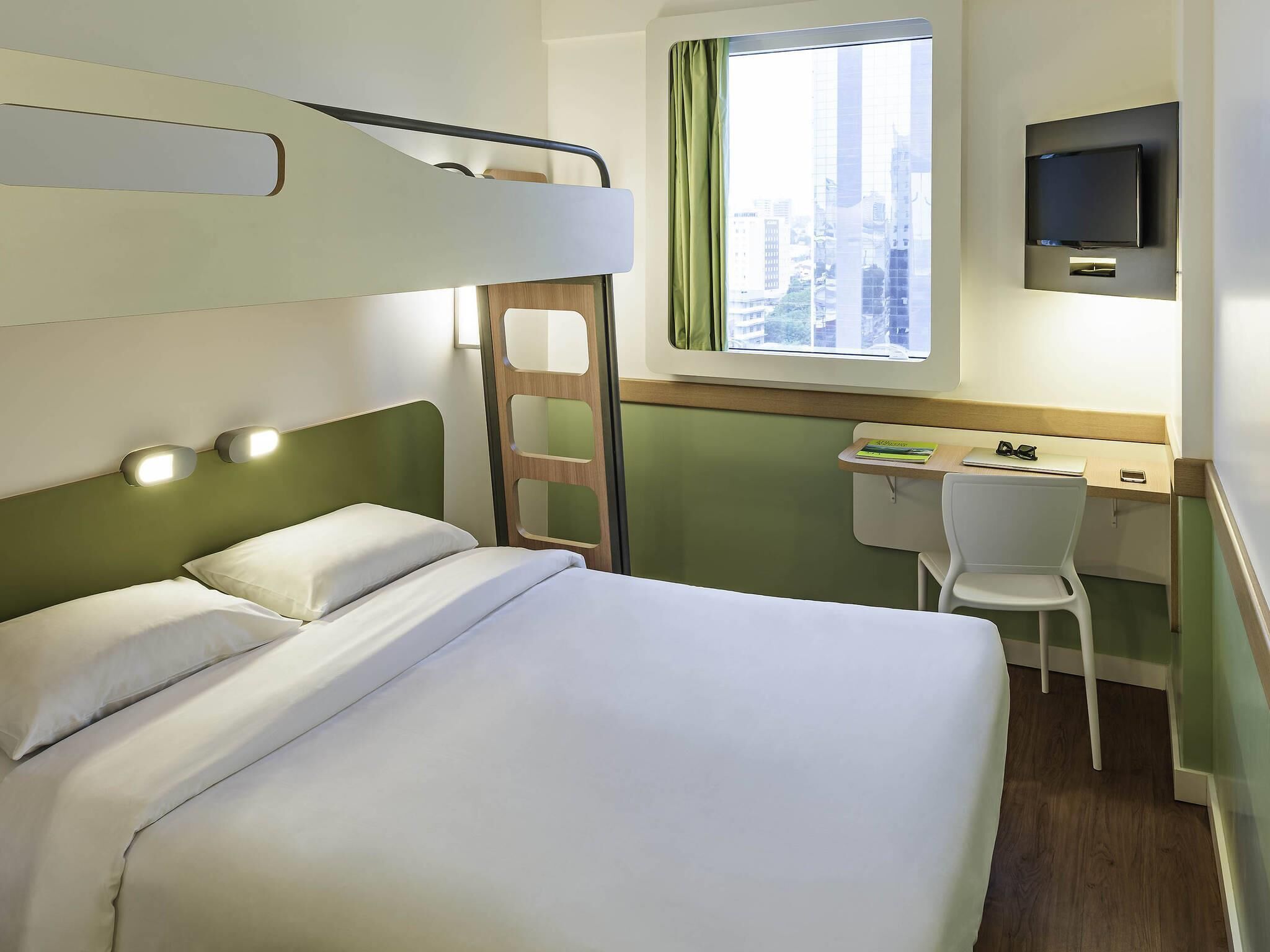 Un lit superposé avec une échelle, un bureau avec une chaise, un téléviseur fixé au mur et une fenêtre avec un rideau.