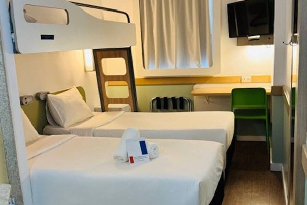 A compact hotel room with two beds, a small kitchenette, a television, and a window with curtains.