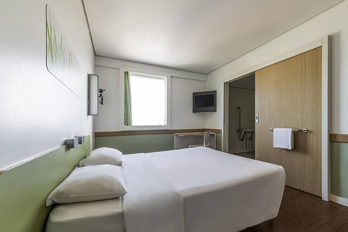 A hotel room with a bed, a TV, a window with curtains, a door, and a towel rack.